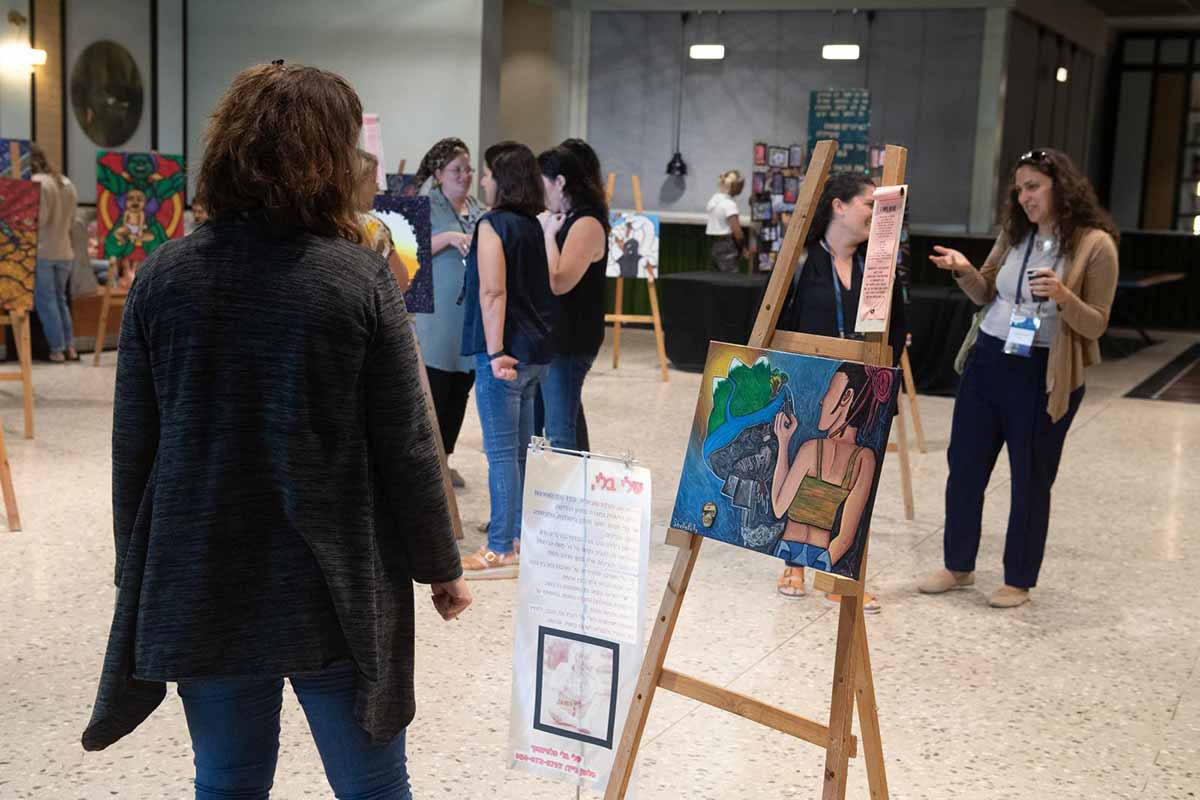 עשור לענבלים - תערוכת ציורים בודדים על חצובות מעץ. באולם עומדות משתתפות באירוע.