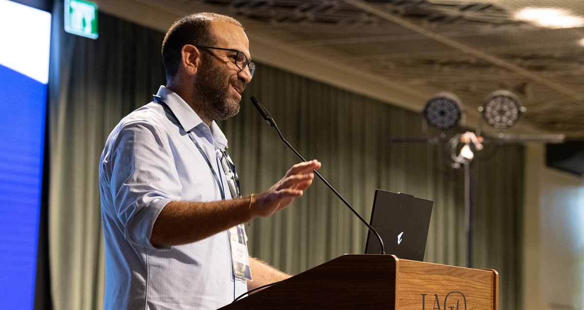 אבישי פריזדה עומד ליד דוכן נואמים מעץ, מיקרופון מהדוכן מגיע קרוב לפיו, ומחשב על הדוכן. על הדוכן כתוב באנגלית: LAGO.