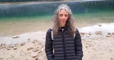 שירה טל עומדת על שפת נהר, לבושה במעיל שחור.