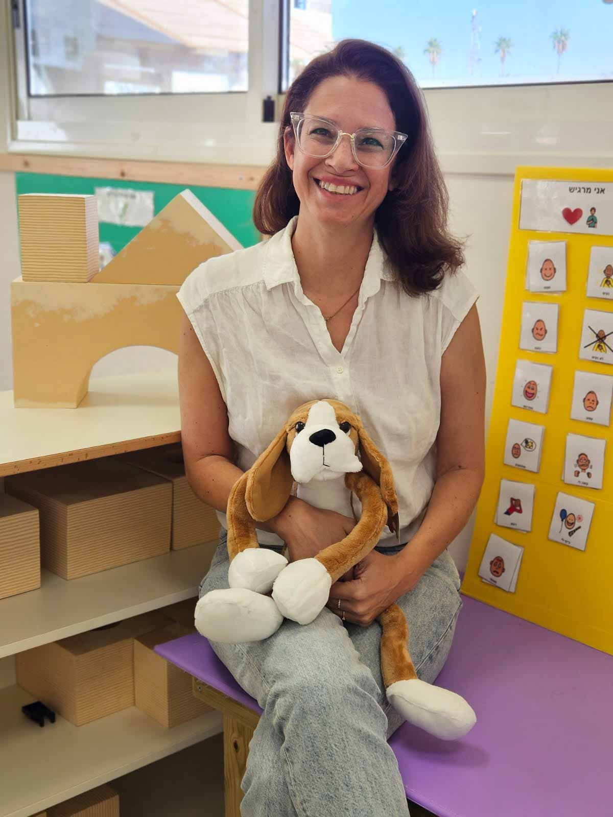 ענבר מרום עם חיבוקי יושבת על ספסל בפינה ישיבה. מימינה לוח עם תמונות. משמאלה מדפים עם קופסאות עץ. ברקע שני חלונות.