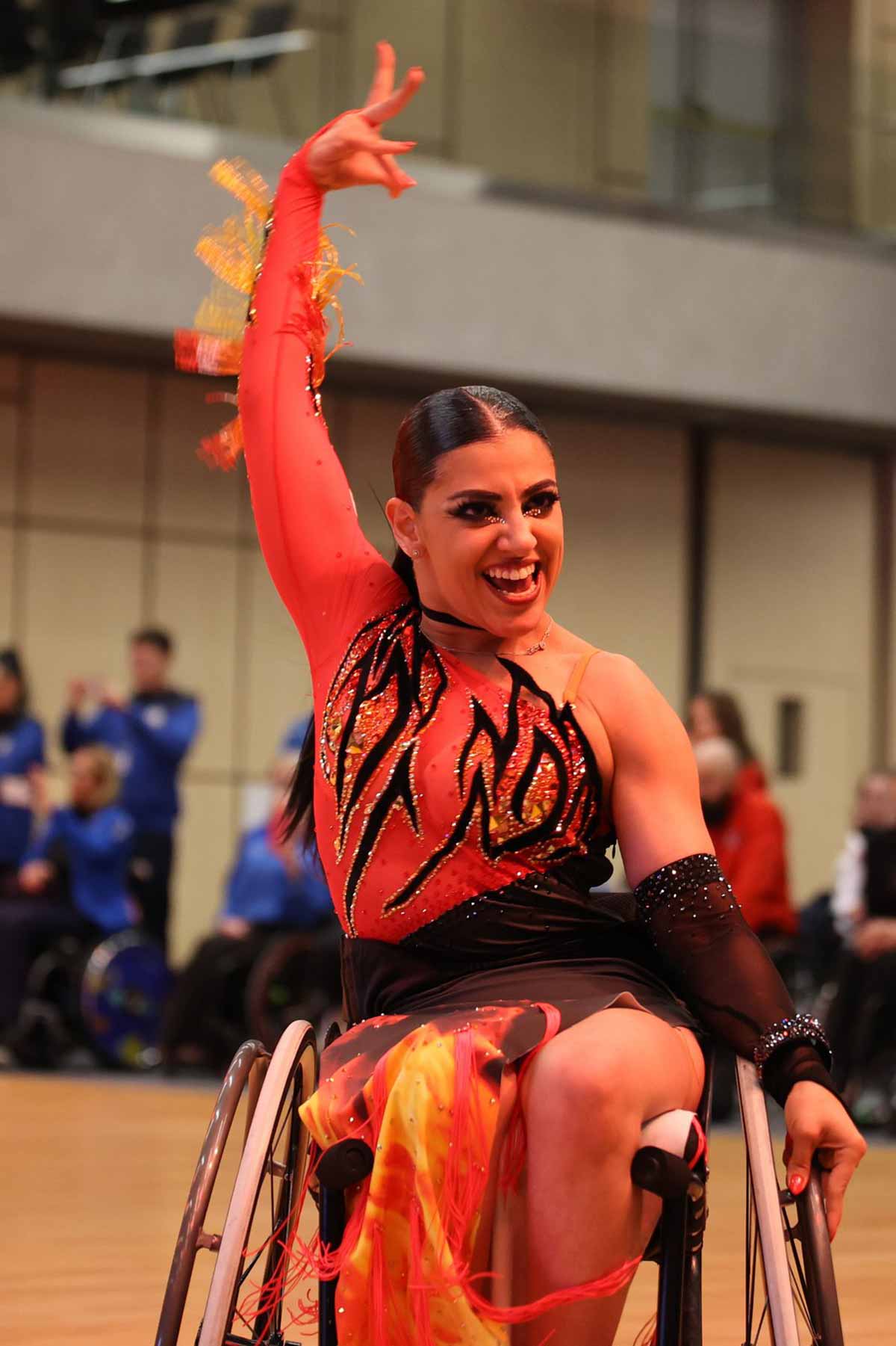 תומר מרגלית אלופה אירופה בריקודים בכסאות גלגלים. לבושה בשמלה אדומה ופסים שחורים יושבת ומניפה את ידה הימנית למעלה.