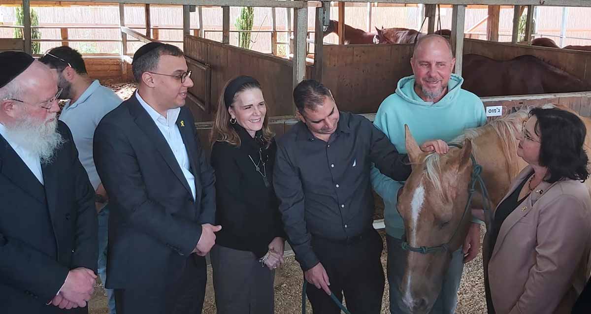 אורוות סוסים. חמישה אנשים מתבוננים בסוס ולידו אישה. ברקע סוסים באורווה.
