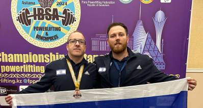 שי רוזין עם המאמן האישי דניאל ריבקין אוחזים בדגל ישראל. על צוארו של שי המדליה של IBSA. מאחוריהם הסמל של IBSA באנגלית עם הכיתוב: Astana 2024, POWERLIFTING & BENCHPRESS