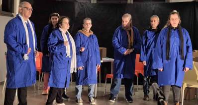 הצגה של קבוצת התיאטרון של אקים חדרה. שבעה שחקנים ושחקניות בבגד כחול ועם צעיפים לבנים, שחורים וחומים על הבמה. מאחוריהם כסאות.