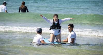 מרכז פעילות חדש בחוף גורדון לצעירים עם צרכים מיוחדים ואוכלוסיות שעברו טראומה. ילדה עם הברכיים על גלשן, ווידיים מונפות לצדדים, ושניים תומכים בגלשן.