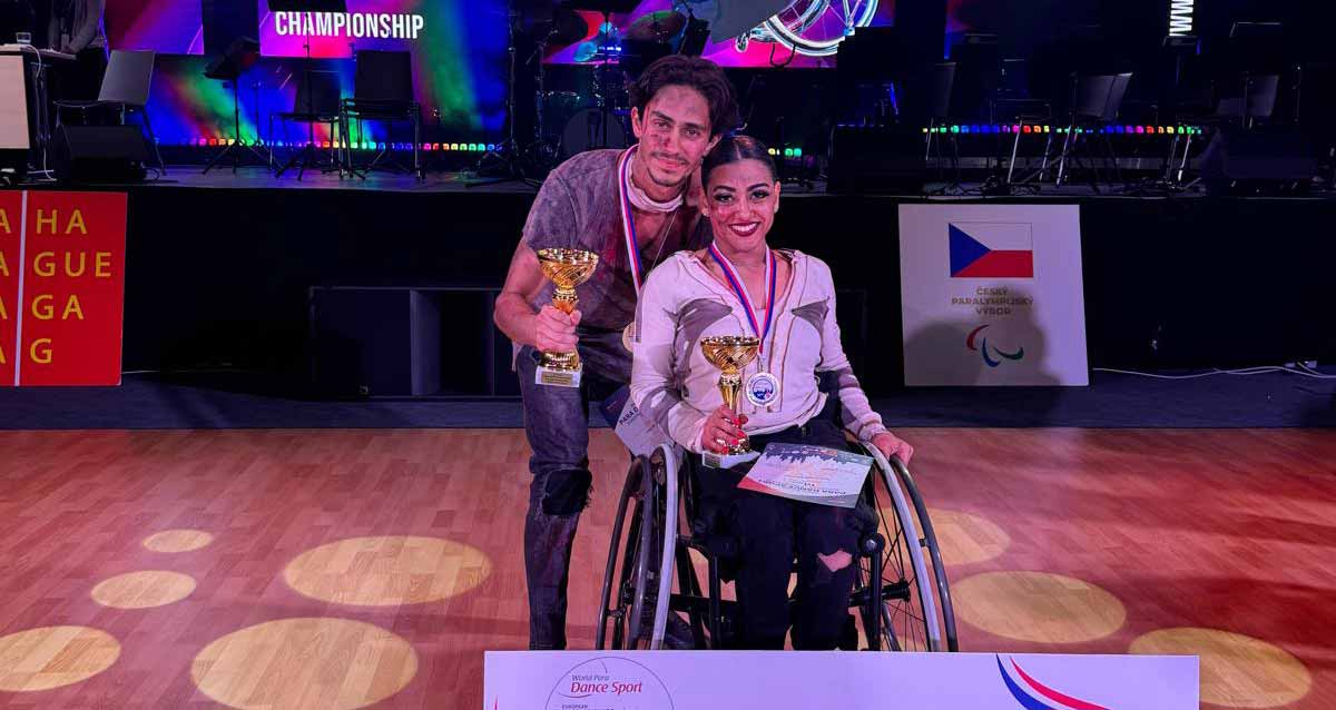 אלופי אירופה בריקוד בכסאות גלגלים עם תלמידי קריית ים. תמונה של רקדנית בכיסא גלגלים עם גביע ביד ומדליה על הצואר. מאחוריה עומד בחור עם גביע ומדליה על הצואר.