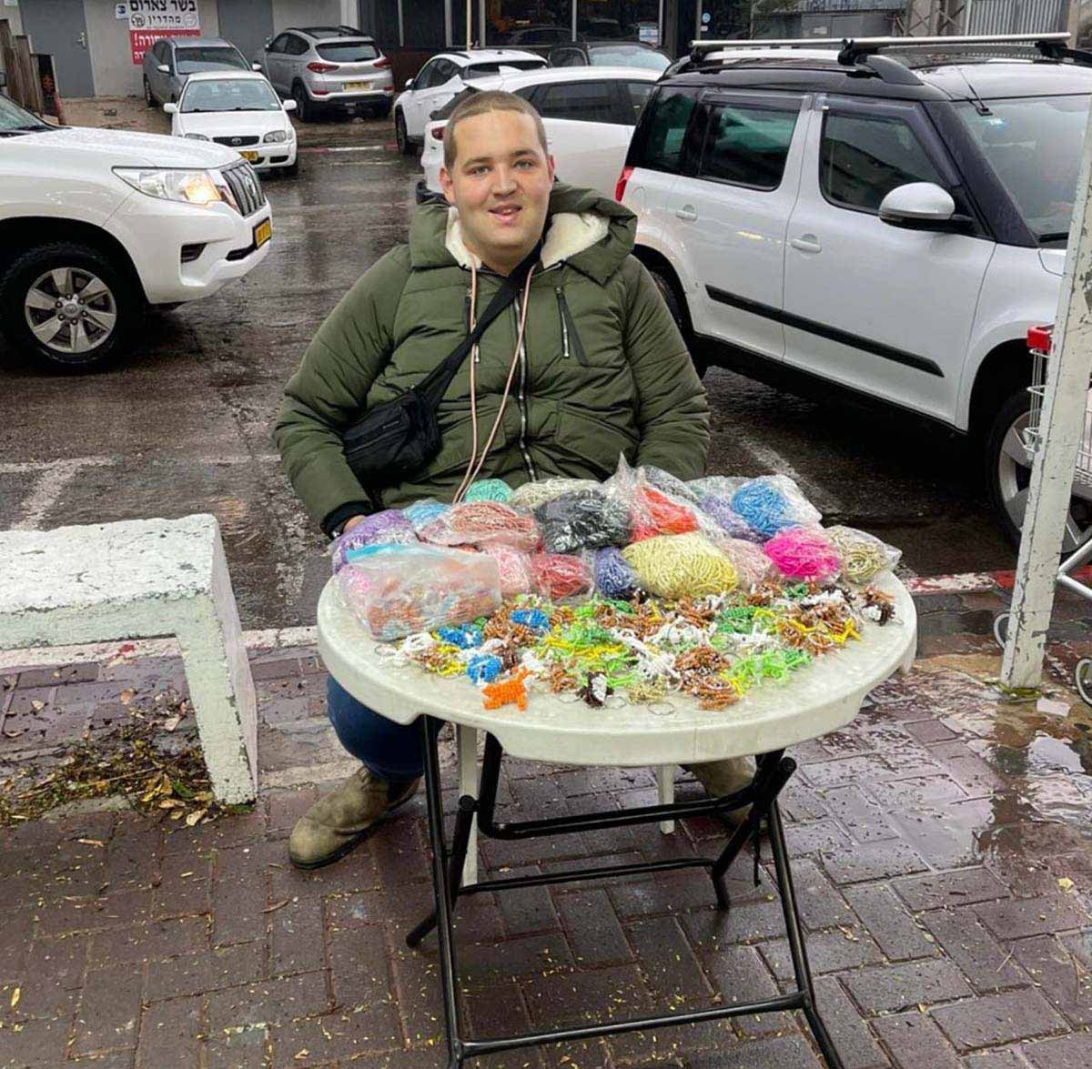 החרוזים של איתן. איתן יושב במדרכה ולפניו שולחן עגול עם מוצרים מחרוזים, בתפזורת ובשקיות פלסטיק שקופות. מאחוריו כביש ומכוניות חונות.