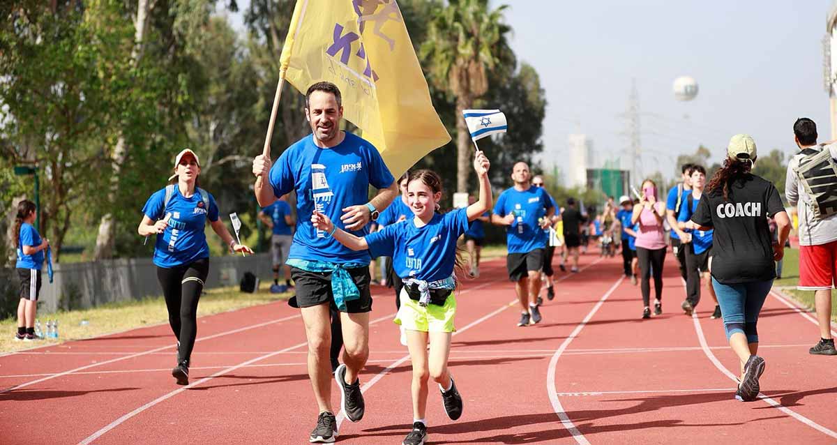 מרוץ איתן RUN על משטח חום. בחזית אב ובתו עם דגל המדינה ודגל צהוב של איתן. אחריהן במסלול רצות ורצים רבים.