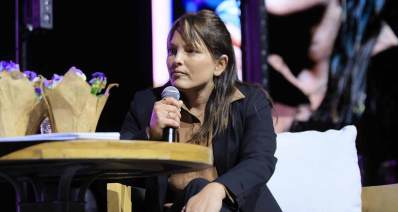 שורדת השבי עמית סוסנה יושבת ומיקרופון בידה. על השולחן לפניה שני עציצים עם פרחים סגולים.