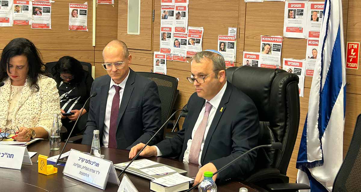 ח"כ גלעד קריב יושב על כסא מנהלים. מאחוריו על הקיר תמונות של חטופים. לפניו שלט עם הכיתוב: חה"כ גלעד קריב, יו"ר ועדת העליה, הקליטה והתפוצות. לימינו יושבים עוד שני חברי הוועדה.