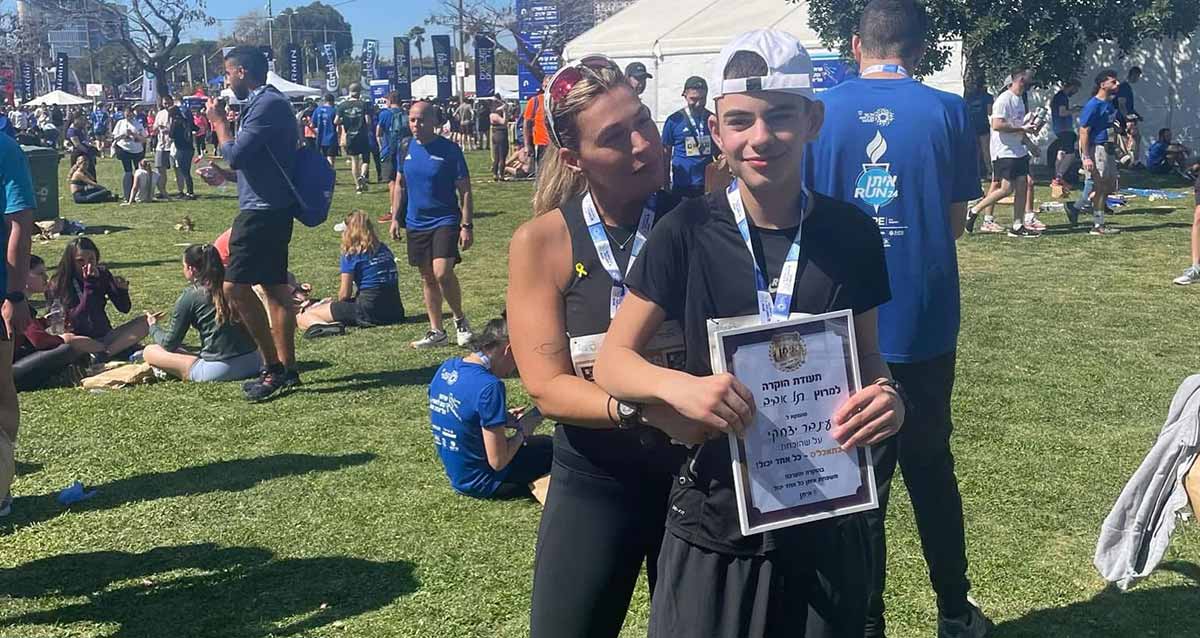 עינבר יצחקי בן 13.5 ואימו מירב במרוץ תל אביב. עינבר עם תעודת הוקרה. ברקע ספורטאים על דשא.