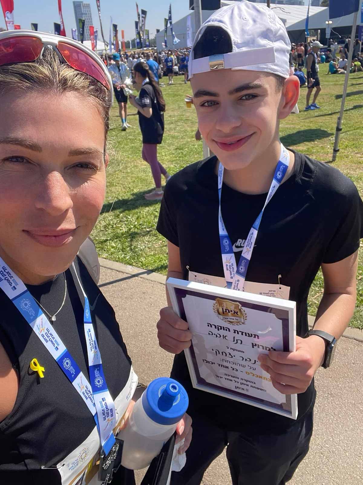 עינבר יצחקי בן 13.5 ואימו מירב במרוץ תל אביב. עינבר עם תעודת הוקרה. ברקע ספורטאים על דשא.