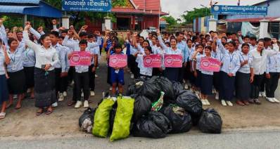 קמבודיה - ניקוי הסביבה. קבוצה גדולה של ילדות, ילדים, נערות ונערים בחולצות תכלת עם שלטים ורודים וכיתוב באנגלית: GOOD DEEDS DAY. לפניהם ערימה גדולה של שקיות שחורות וצהובות עם אשפה.