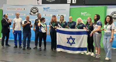 נבחרת הנשים בכדורשער, ברלין 2025 בצילום קבוצתי עם דגל ישראל. מאחור על הקיר שלט בד גדול ולמעלה הכיתוב באנגלית: SPORTMETROPOLE BERLIN. וסמלים על השלט.